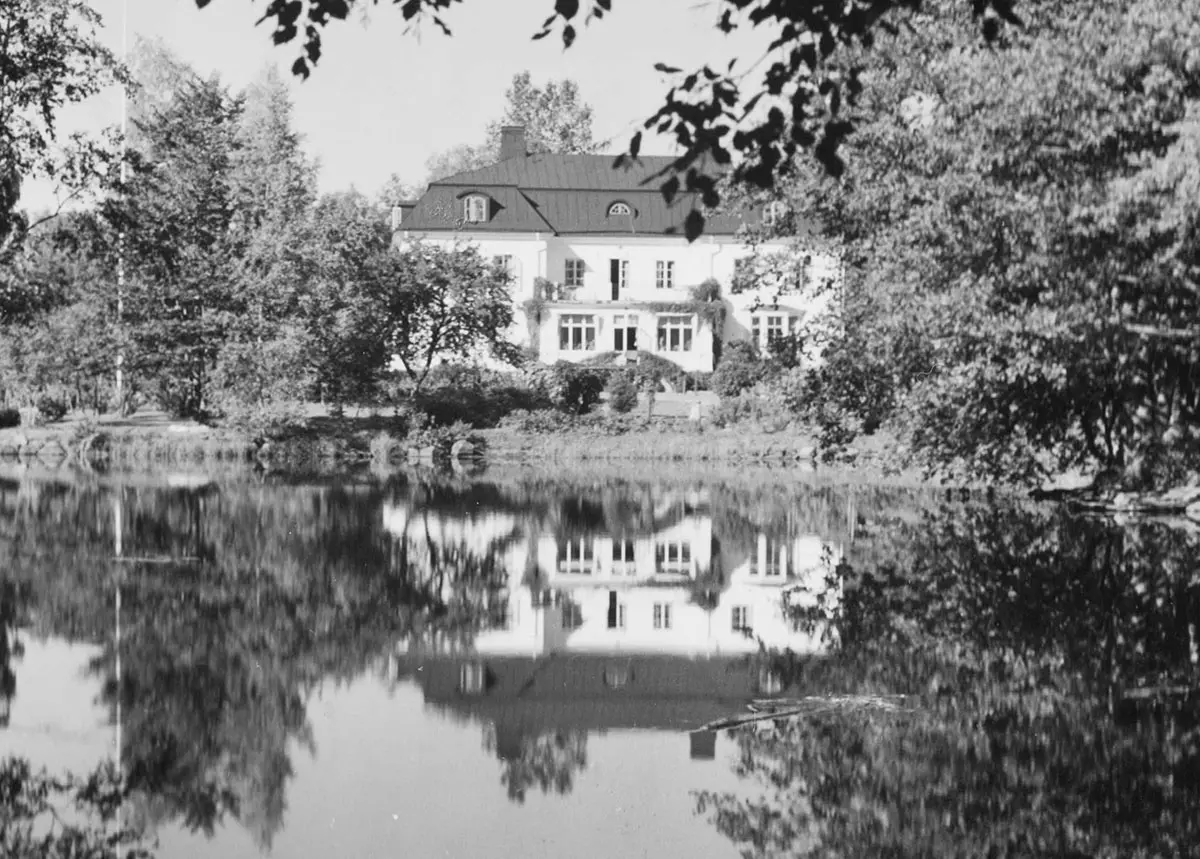En pampig vit villa speglas i ån med lummiga träd på båda sidor.