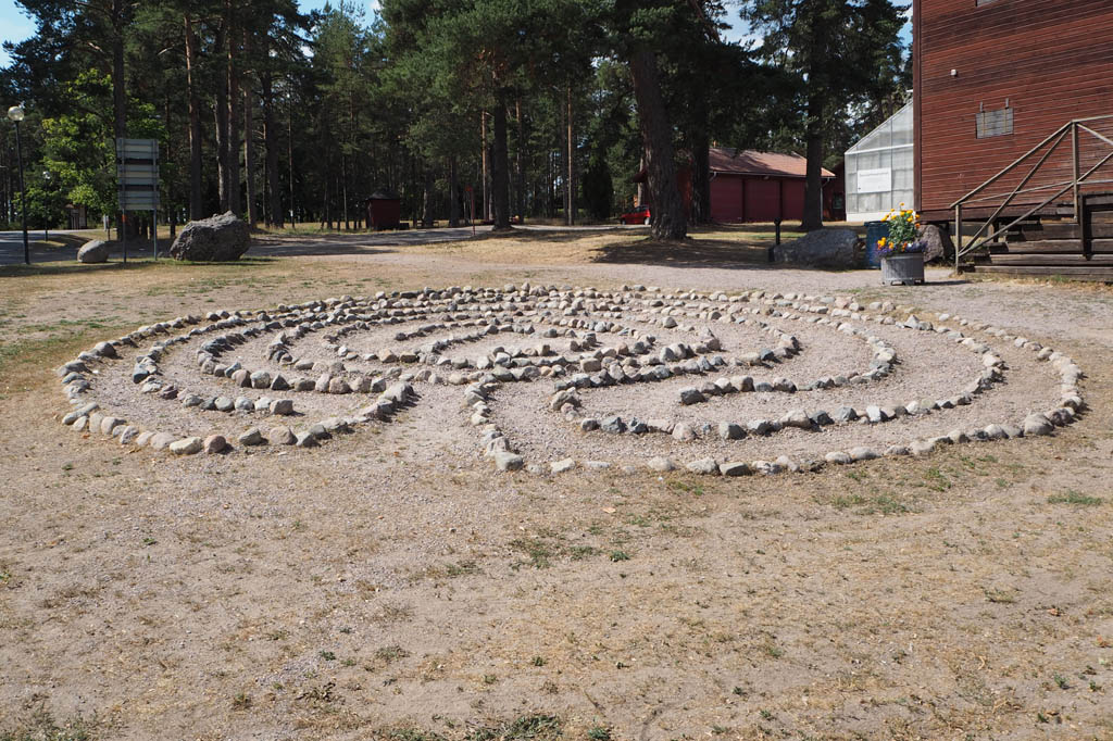 En rund labyrint, formad av rader av knytnävsstora stenar, på en slänt utanför en röd timrad byggnad. 