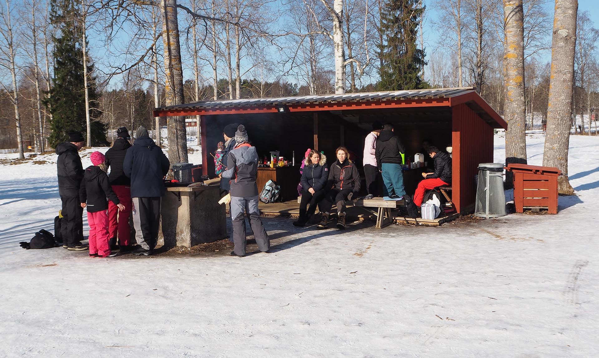 Ett tiotal barn och vuxna tar en paus i ett rött vindskydd vid en grillplats.