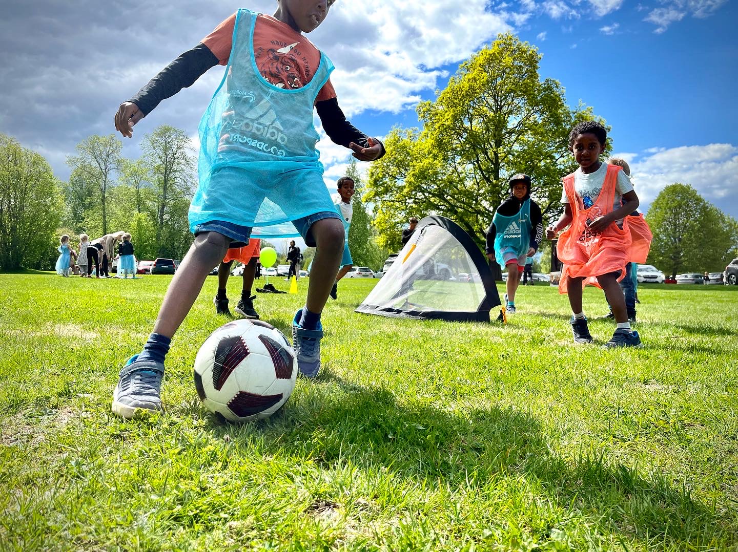 Barn i färgglada västar spelar fotboll på en gräsplan utomhus.
