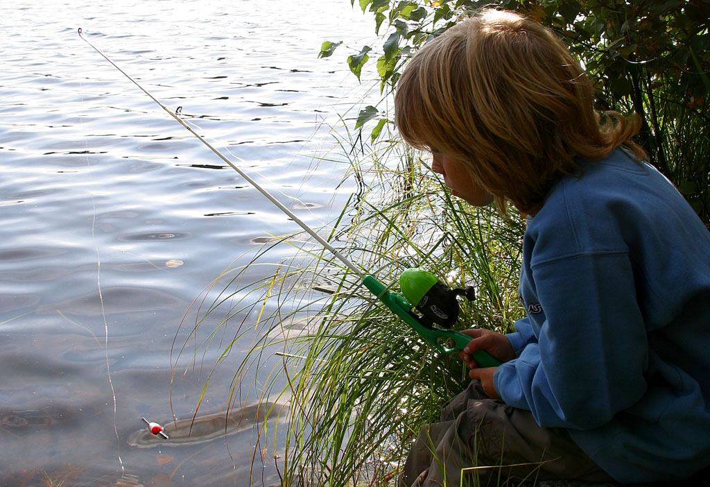 Ett barn siiter vid kanten av en sjö med ett fiskespö i handen.
