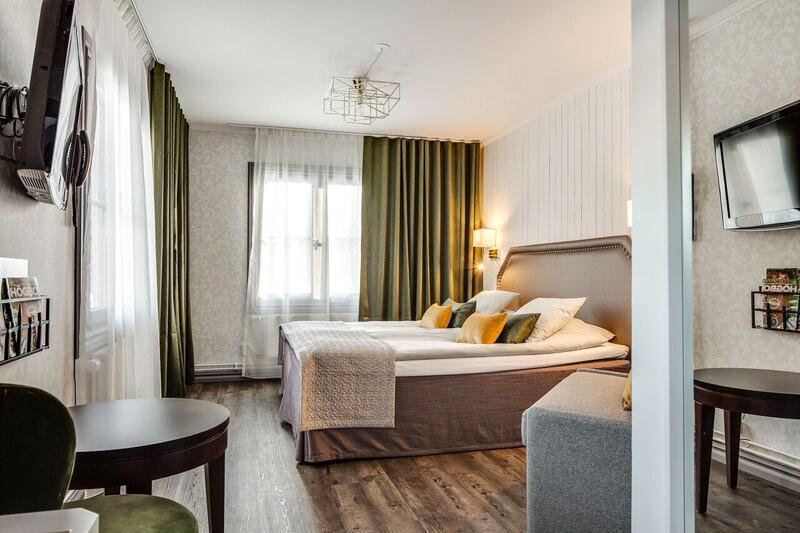 Hotellrum med dubbelsäng, två stora fönster, ljusa väggar och trämönstrat golv.