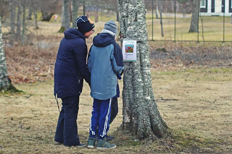 En förälder och två barn går sportlovets tipsrunda tillsammans. Dom läser en fråga som sitter fäst på ett träd i bruksparken. 