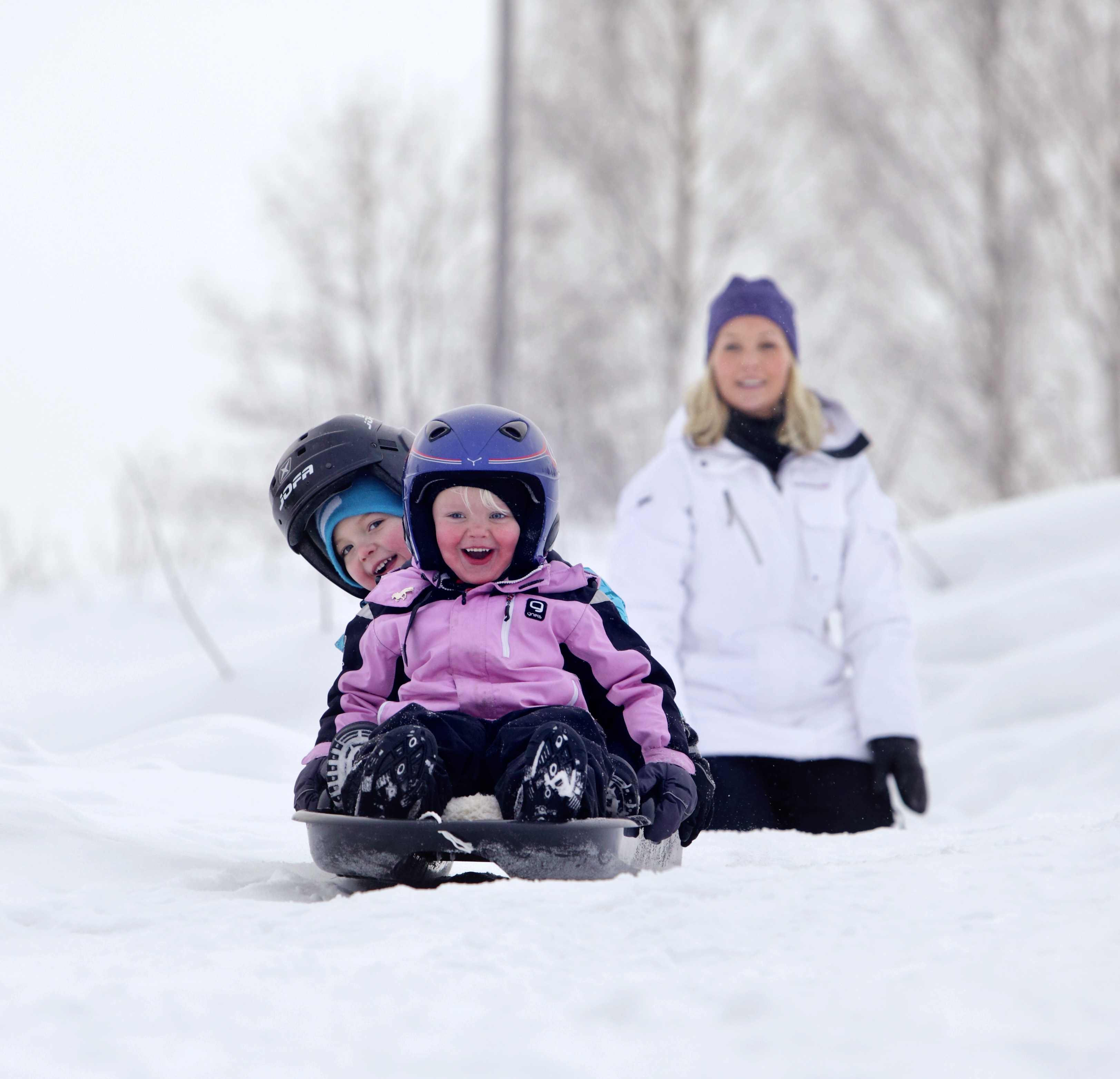 Två vinterklädda små barn åker pulka nerför en stor snöig backe.